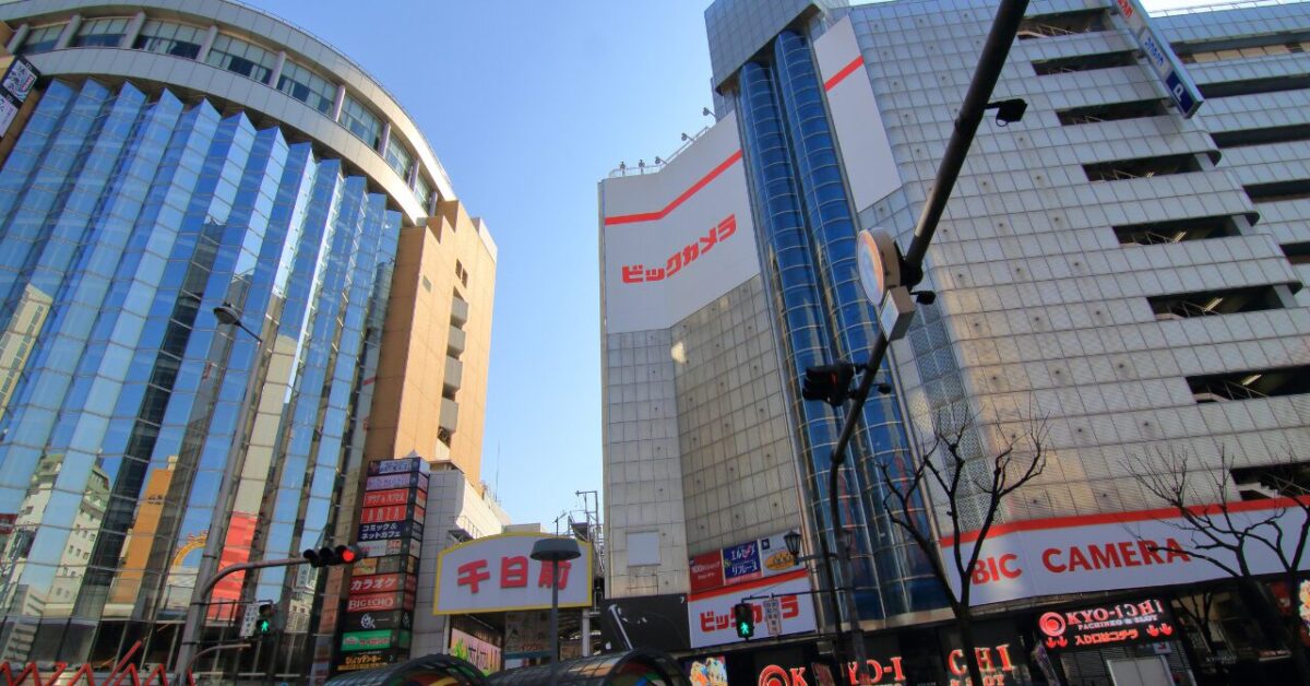 大阪・千日前エリアの街並み。中央にビックカメラなんば店の外観が見え、周囲にはガラス張りの高層ビルや商業施設が立ち並ぶ。晴天のもと、多くの看板が並ぶ賑やかな都市風景。